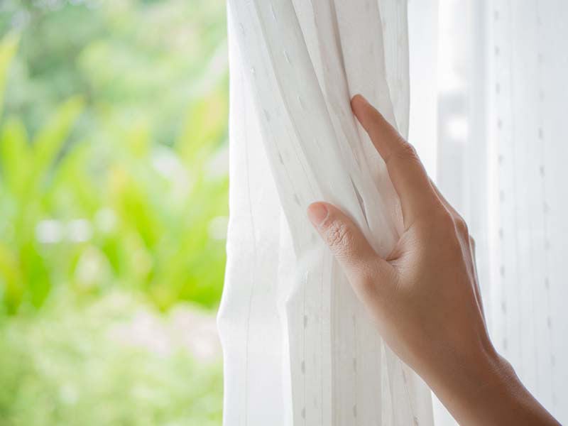Woman's hand opening curtains in the bedroom with natural light and garden background.