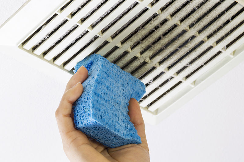 Close up horizontal photo of female hand cleaning dirty bathroom fan vent cover with blue sponge