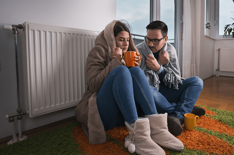Two people sit next to each other, warming up by a radiator. When Should You Replace Your Furnace?