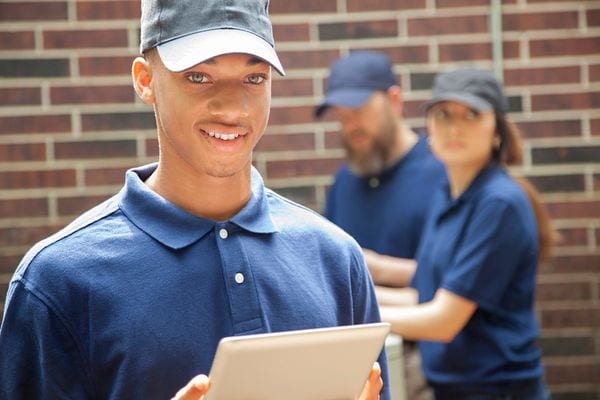 Young HVAC technician smiling holding clipboard with coworkers in background.